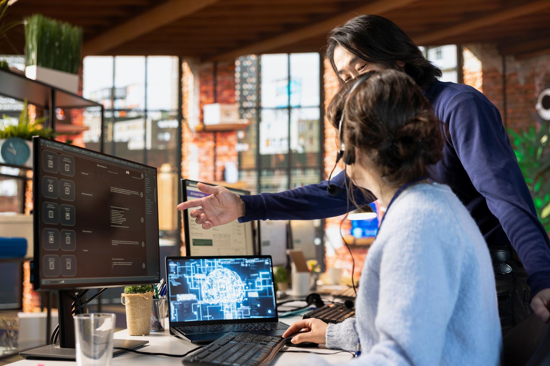 Call center manager guiding intern on how to use AI chatbot at office desk on PC